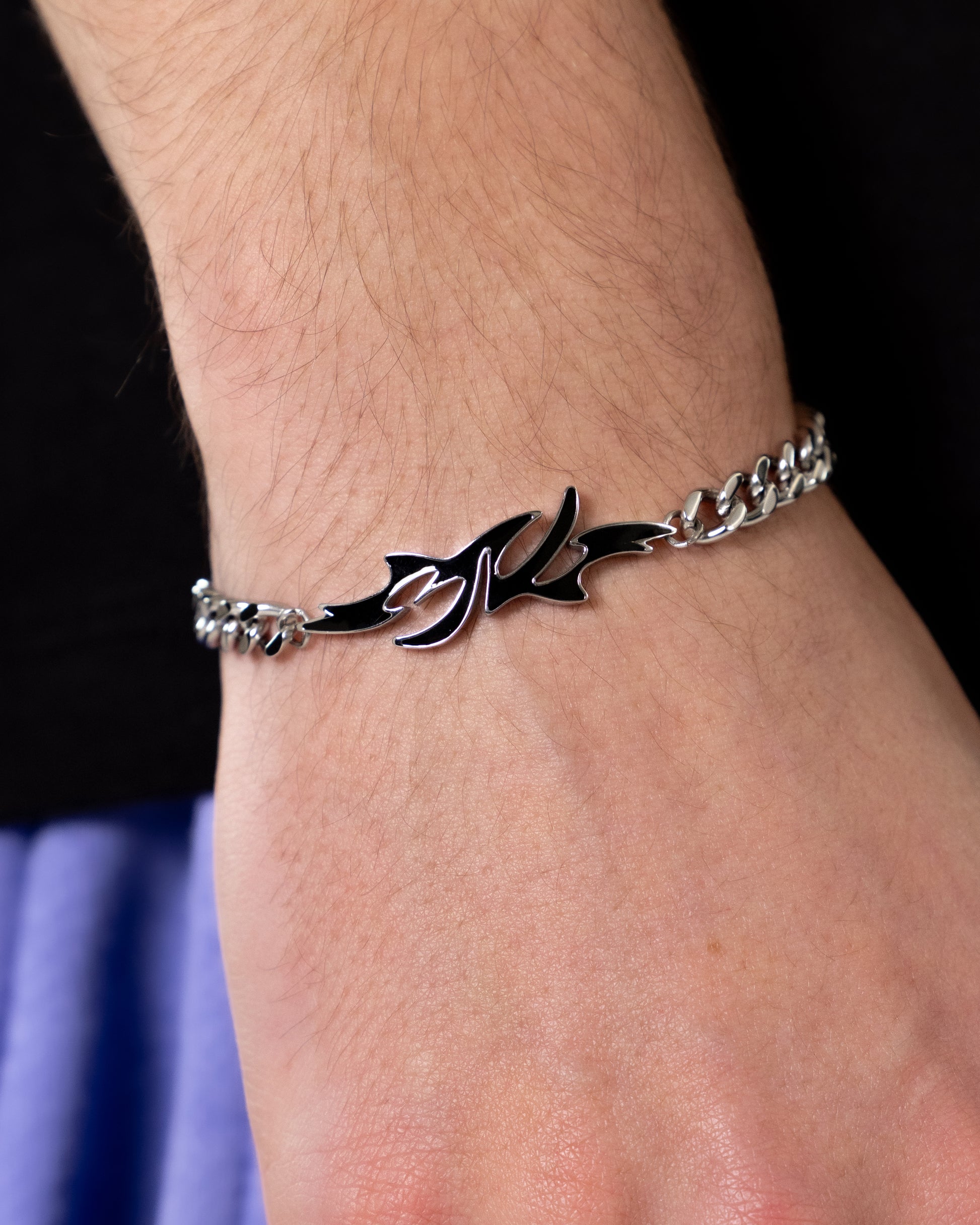 Das Armband liegt eng um das Handgelenk und zeigt mittig ein flammenförmiges „REVED“-Logo aus poliertem Metall mit geschwungener, asymmetrischer Silhouette.