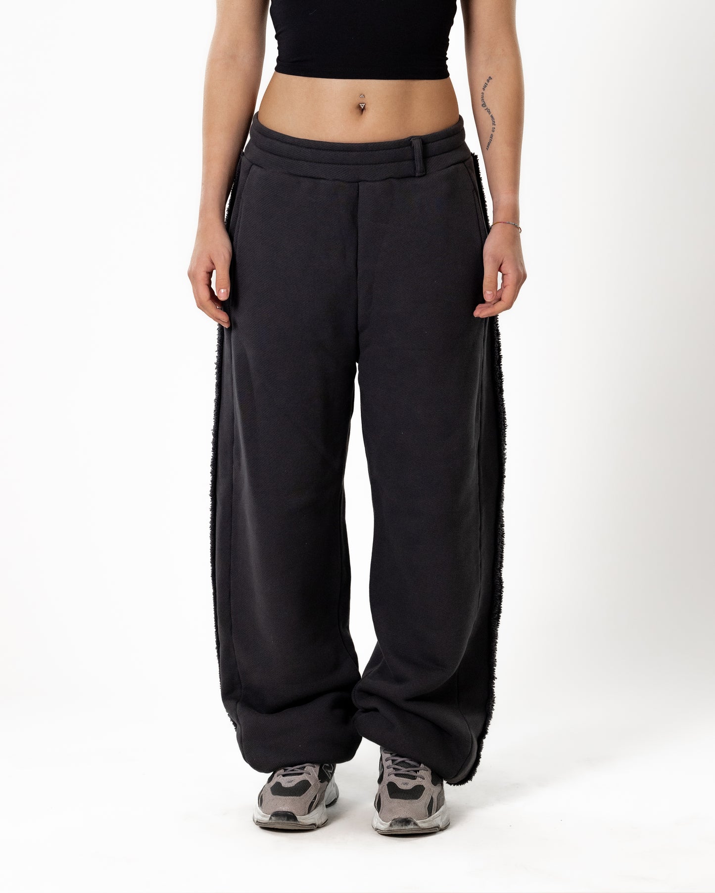 Frontansicht einer schwarzen Jogginghose mit weitem Bein, breitem Taillenbund und seitlicher Fransenkante, getragen von einer Person mit schwarzem Crop-Top und Sneakers.