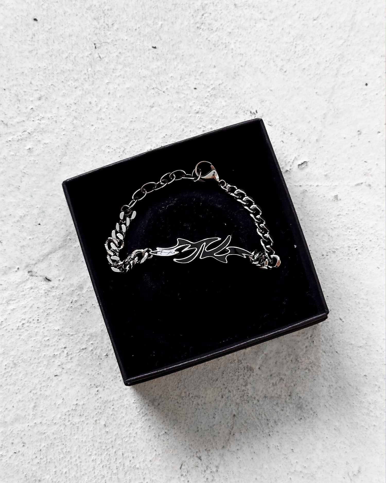 Das flammenförmige Logo zentriert in schwarzer Schmuckbox, mit geöffnetem Karabinerverschluss und feiner Gliederkette sichtbar am Rand.